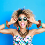 Black woman with curly hair and sunglasses against a blue background smiles because she is excited for summer