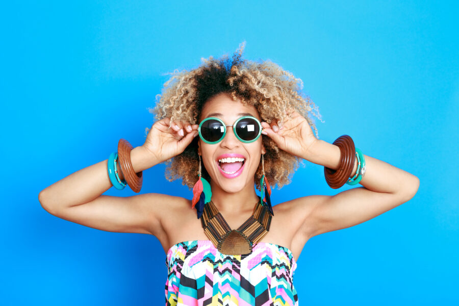 Black woman with curly hair and sunglasses against a blue background smiles because she is excited for summer, dentist in bentonville