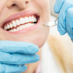 Closeup of a woman smiling as blue gloved hands hold a special dental mirror to examine her teeth during a routine checkup