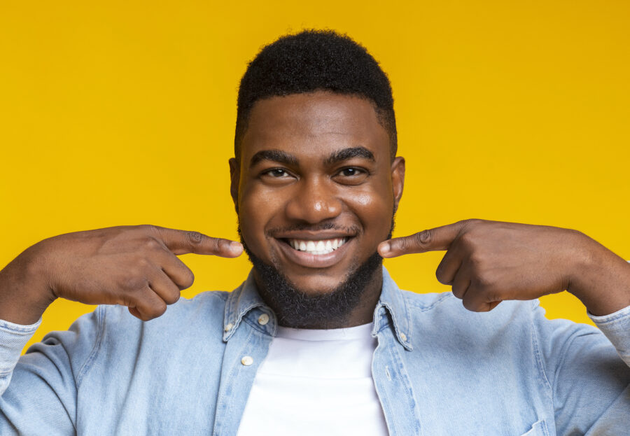 Black man against a yellow wall smiles and points to his teeth after professional teeth whitening