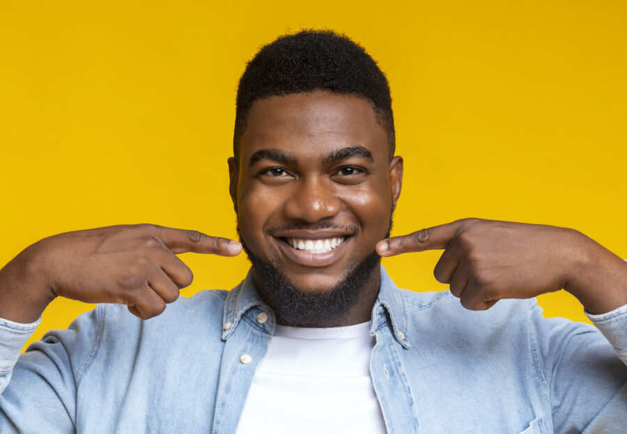 Black man against a yellow wall smiles and points to his teeth after professional teeth whitening, dentist in bentonville