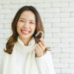 Asian woman in a cream turtleneck smiles as she holds up her Invisalign aligners against a white brick wall