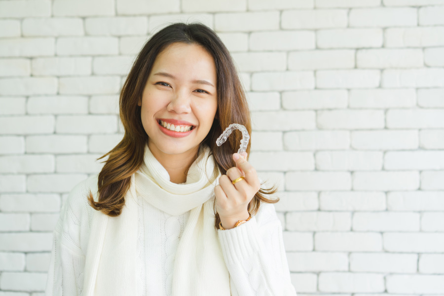 Asian woman in a cream turtleneck smiles as she holds up her Invisalign aligners against a white brick wall, dentist in bentonville